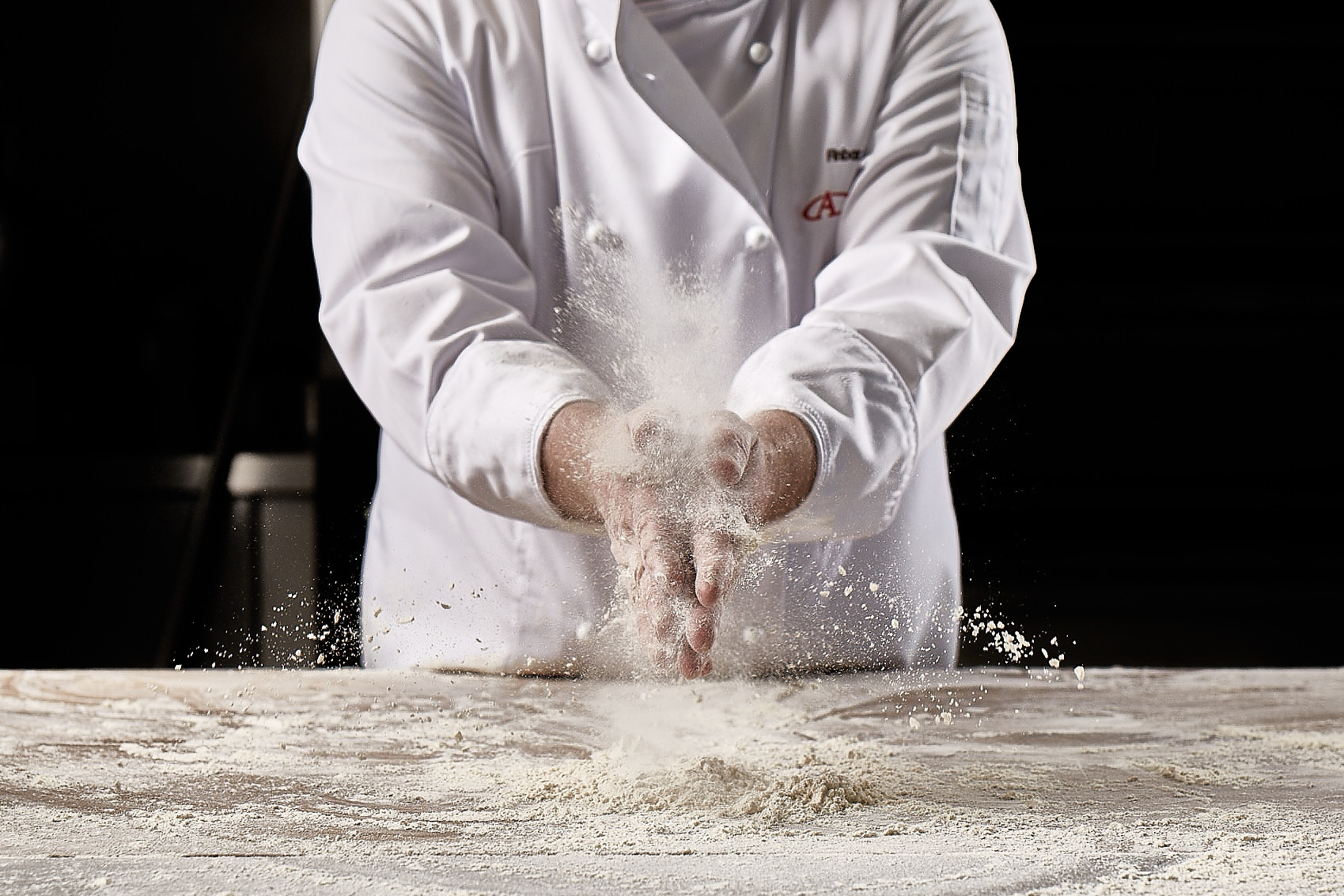 A baker in a white overcoat clapping flour between their hands